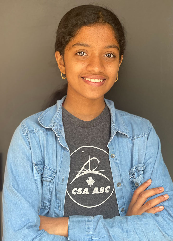 Portrait of Bhavishyaa. Her arms are crossed. She is wearing a grey t-shirt with a Canadian Space Agency logo under a jean blouse.