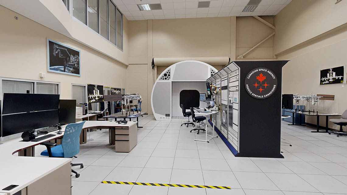 Large training room with various workstations, one of which is mounted to a stylized wall meant to evoke the International Space Station. There are two models of the station in the room, as some photos of station components on the walls.