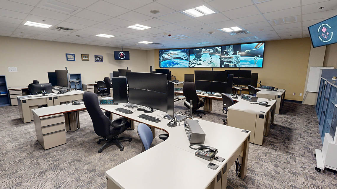 Office area with several workstations. There are three screens at the front of the room to show live views of the ISS.