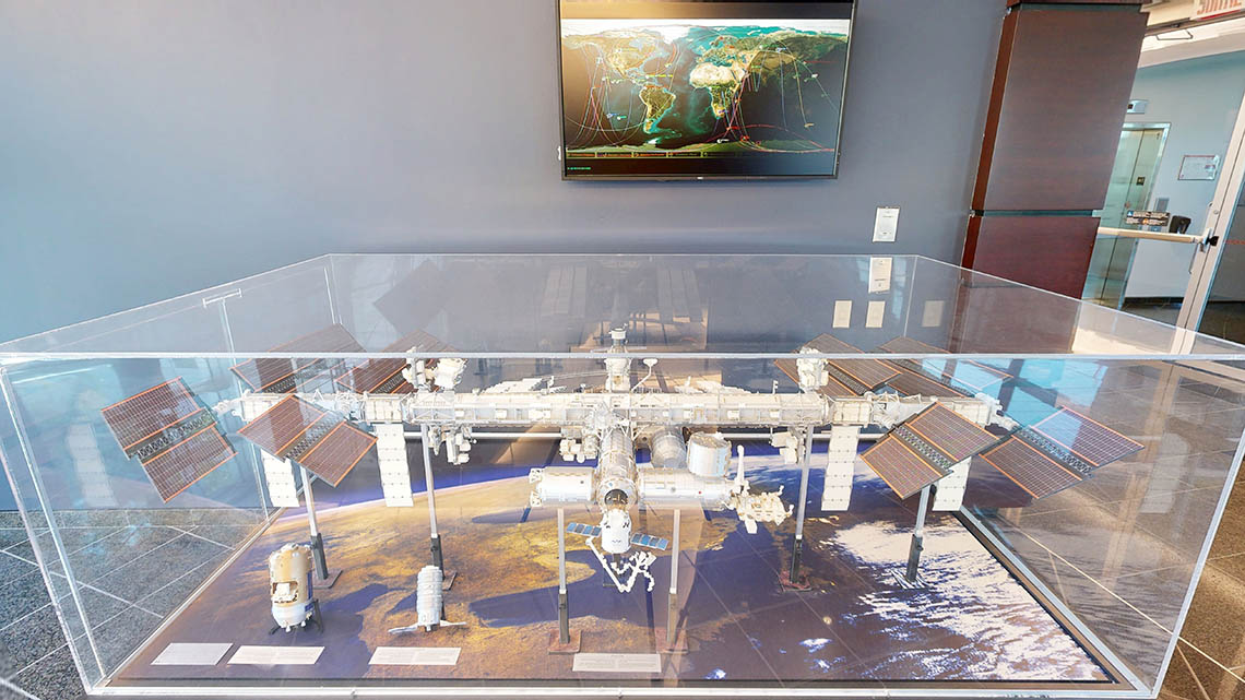 Maquette de la Station spatiale sur un support. Au-dessus, un écran montre le trajet de divers engins spatiaux au-dessus de la Terre.