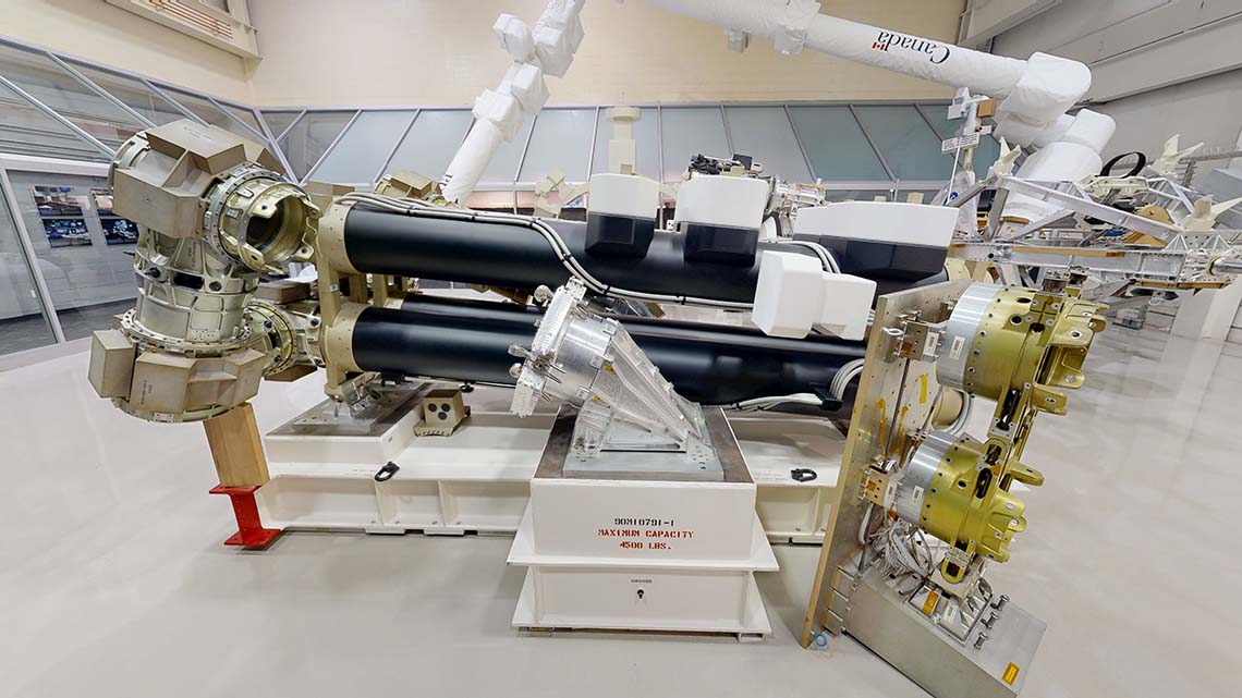 Grand modèle du Canadarm2 plié, sans son capot thermique blanc, avec une structure noire détaillée, incluant des câblages, des joints et des embouts métalliques.