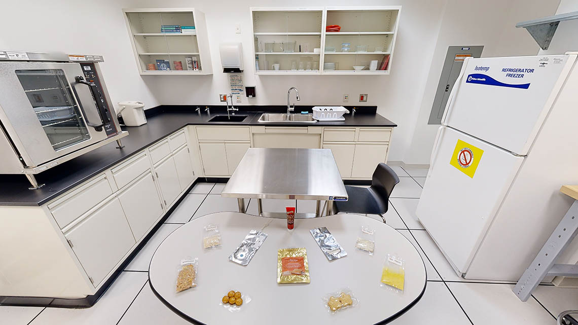 Une cuisine avec divers appareils électroménagers, placards, éviers et un assortiment d'échantillons de nourriture spatiale exposés sur une table.