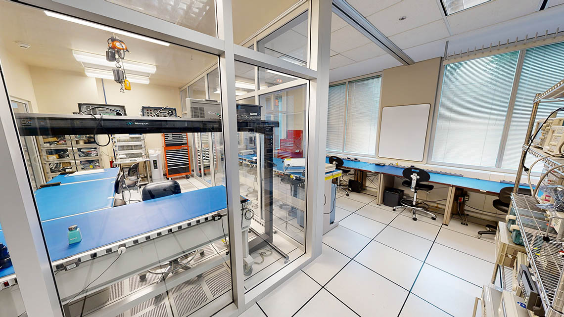 Laboratoire moderne avec des paillasses bleues et chaises noires. La salle blanche est entourée de parois vitrées. Les outils et équipements sont soigneusement rangés.