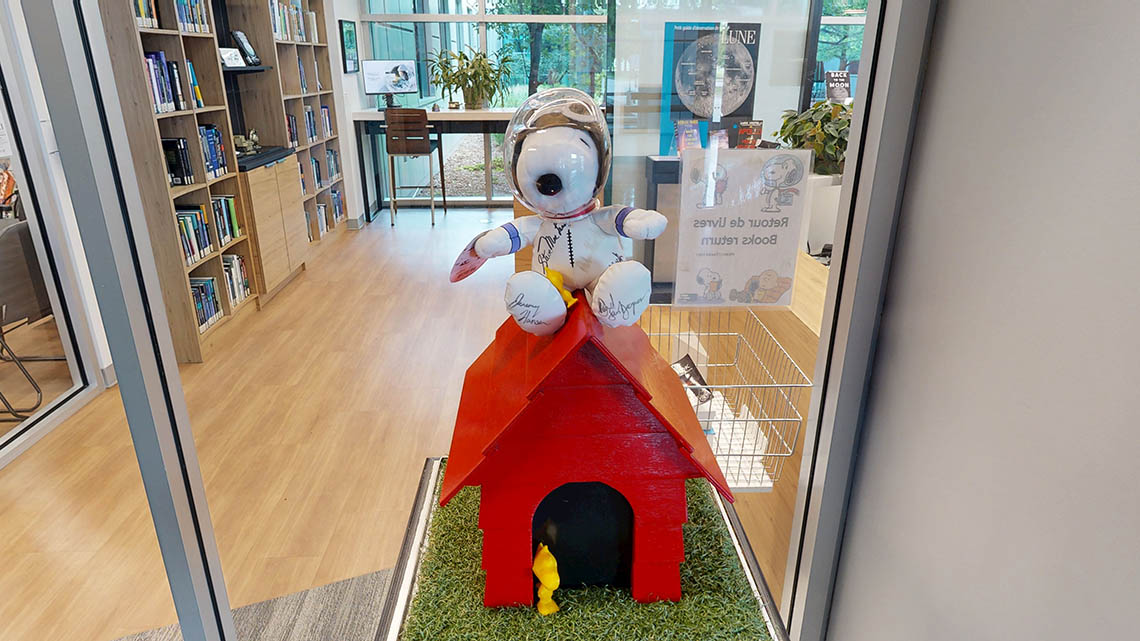A l'entrée de la bibliothèque, un Snoopy en peluche porte un casque d'astronaute et est assis sur sa petite niche rouge, tandis que Woodstock, le petit oiseau jaune, se tient à la porte.