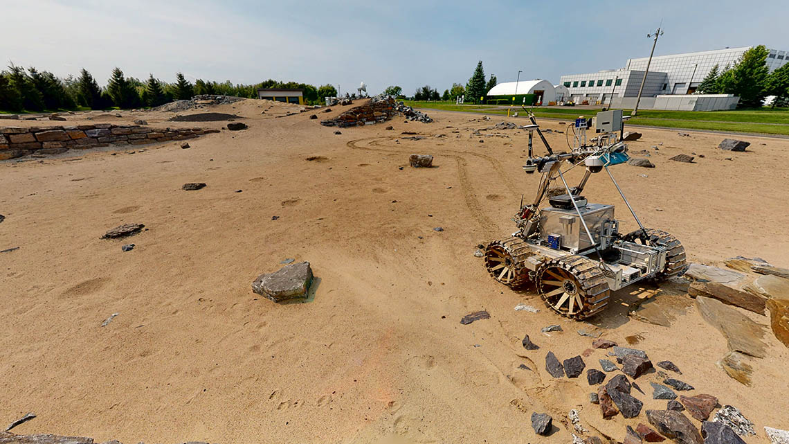 Champ sablonneux avec quelques rochers et collines artificielles. Un rover est visible au premier plan, et des arbres et des bâtiments en l'arrière-plan.