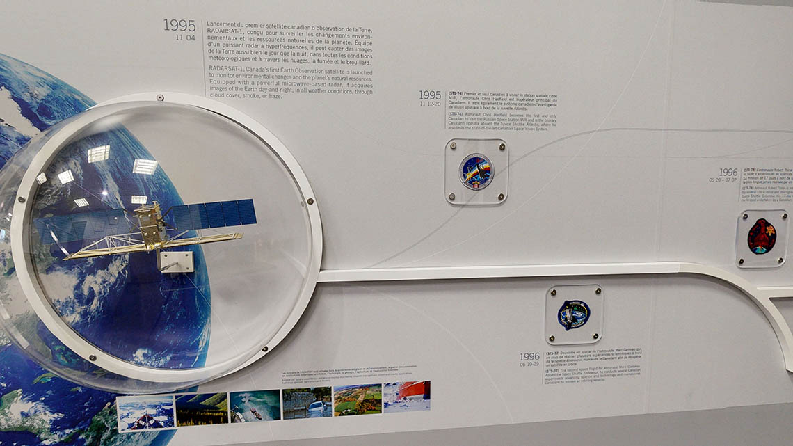 A wall timeline featuring a small model of the RADARSAT-1 satellite, as well as the patches from missions STS-74, STS-77, and STS 78.