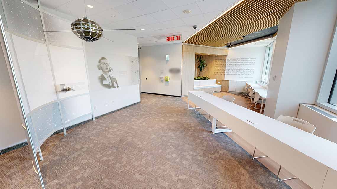 Reception area with desks and seating. A model of the Alouette I satellite hangs from the ceiling. There is text on the wall and a photo of John H. Chapman.