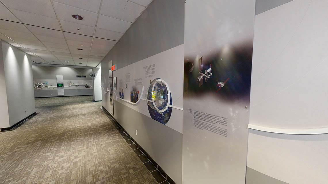 A hallway leading to an open area at the Canadian Space Agency conference centre. A timeline composed of text, images, mission patches, and models runs continuously along the various walls.