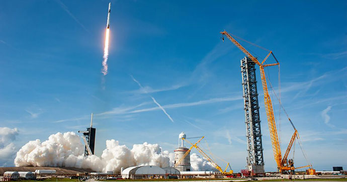 On a sunny day, a Falcon 9 rocket lifts off from a launch pad.