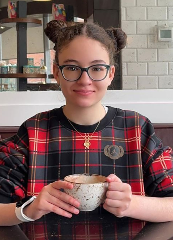Portrait of Maya. She is sitting at a table holding a mug.