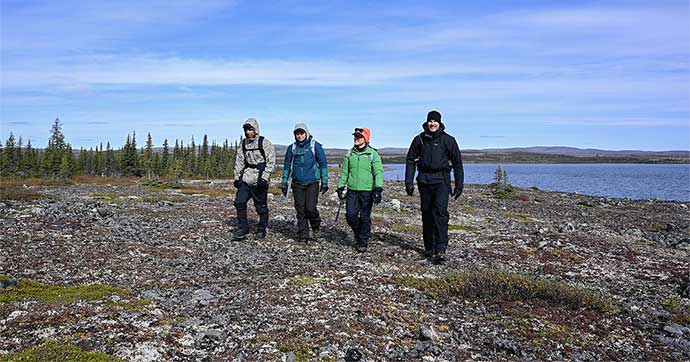 Four people crouch around a large rock. In the background is a body of water.