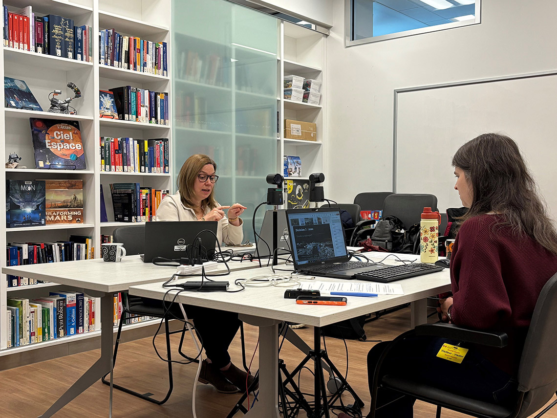 Caroline-Emmanuelle and Olivia are seated across from one another, each with a laptop and webcam in front of her.