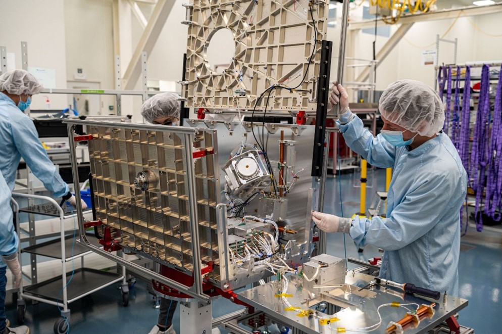 Engineers attach a side panel to a satellite assembly being manufactured in a clean room.