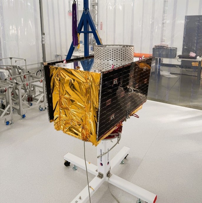 A rectangular satellite with shiny gold wrapping and solar panels on two sides on a stand in a manufacturing facility.