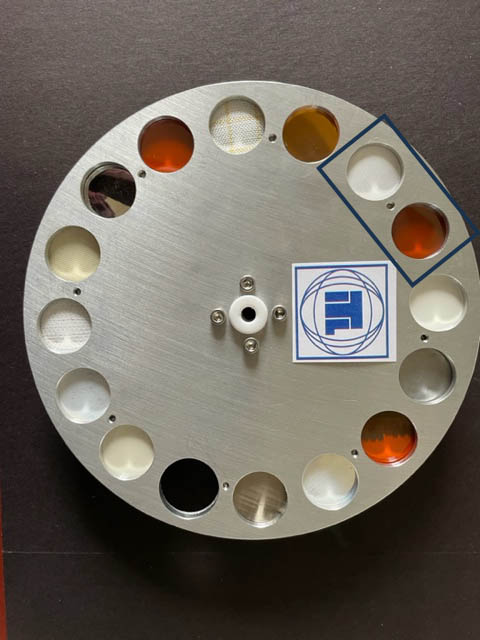 Objet métallique circulaire comprenant plusieurs petits compartiments circulaires, dont certains contiennent des éléments colorés.