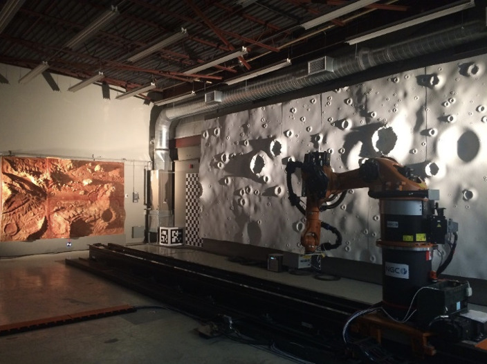 Laboratoire intérieur avec un bras robotique devant un grand mur simulant la surface lunaire.