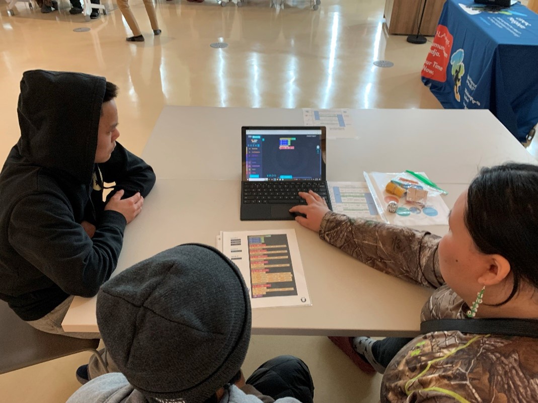 Three students sit at a table with a computer. Instruction sheets are around the computer.