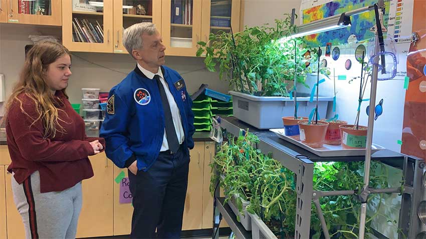 Une fille se tient derrière Robert Thirsk et devant eux se trouve des plants de tomates sur des étagères.