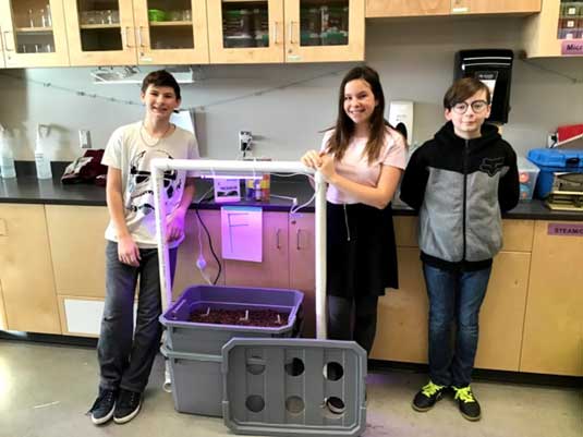 Trois élèves prennent la pose à côté d'un système composé d'un bac et de tubes.
