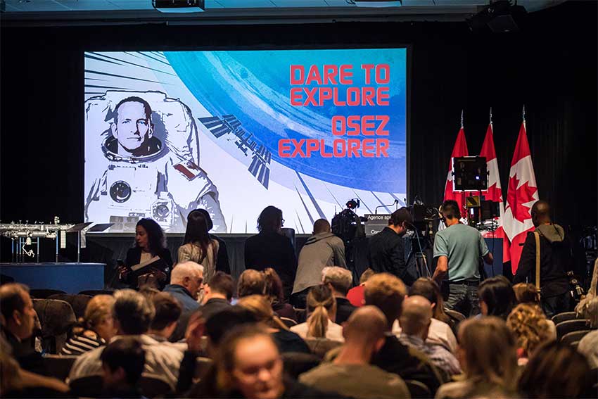 A group of people sitting down in a conference room with reporters standing in front of a stage with a large screen.