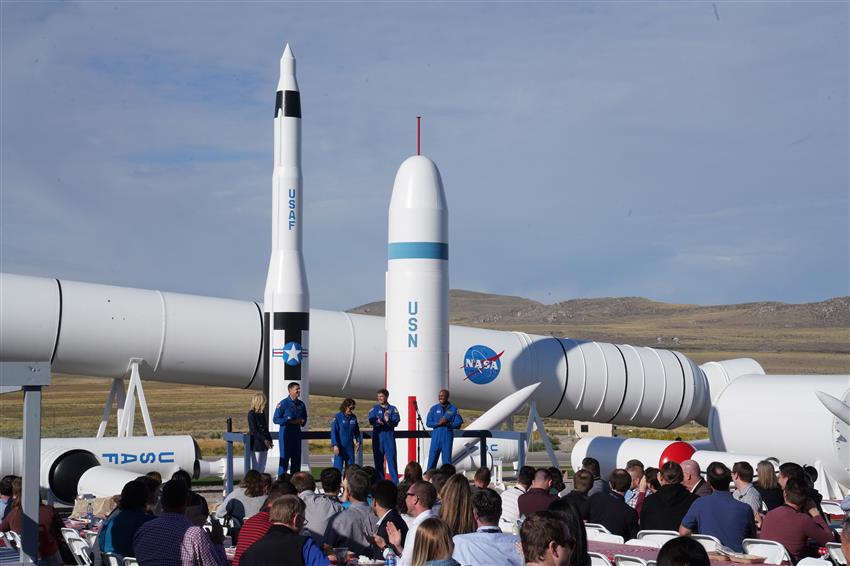 Four astronauts and a woman stand with rockets behind them and an audience in front of them. 