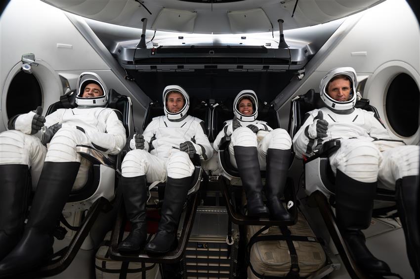 The four astronauts are seated in a mock-up of the Dragon capsule wearing space suits.