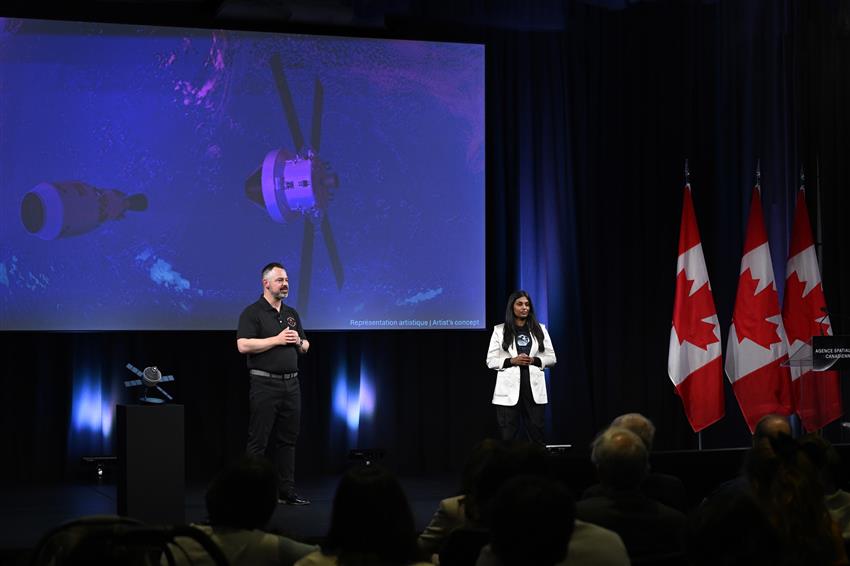 Tim and Kumudu stand on a stage, with a large screen behind them. 