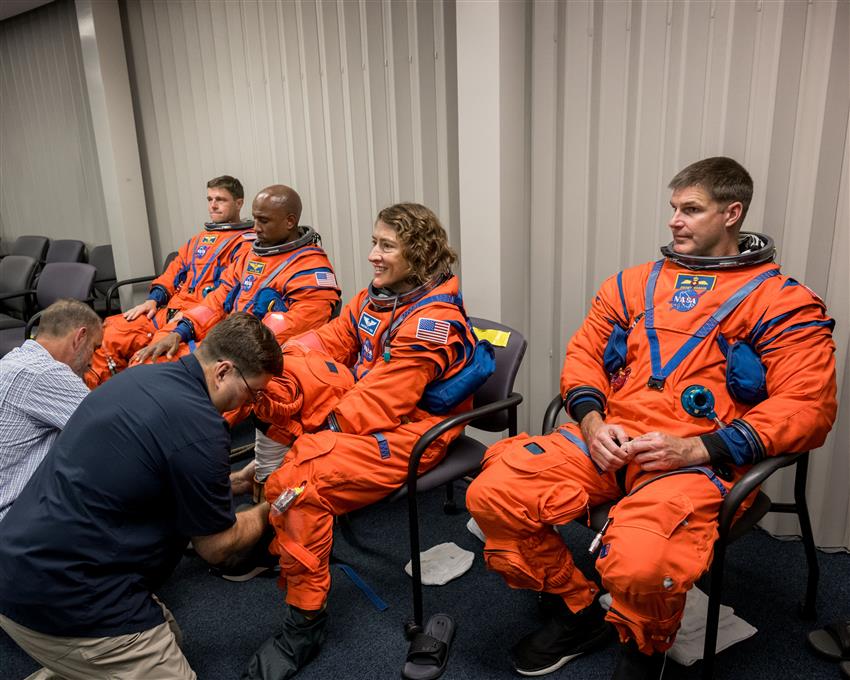 Four astronauts are wearing orange spacesuits, and two people help them put on their boots.
