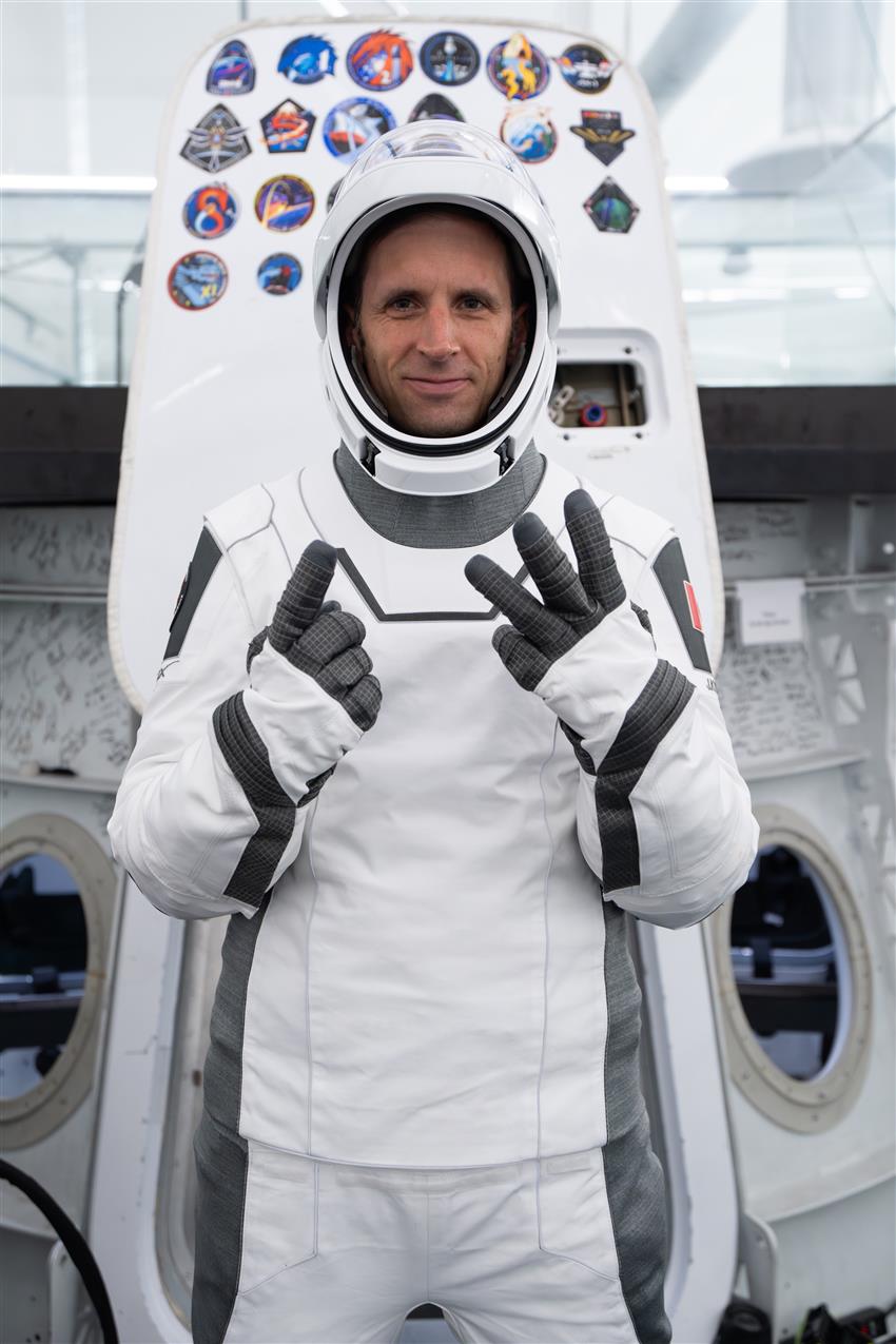 Joshua, vêtu d'une combinaison spatiale de SpaceX, fait le chiffre 13 avec ses doigts.