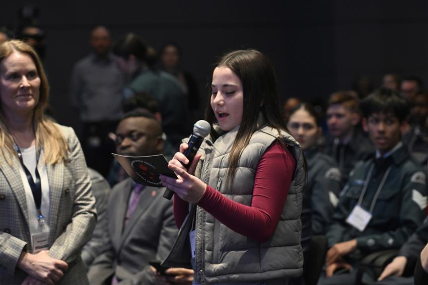 A young girl speaks into a microphone. She is looking at a cue card.