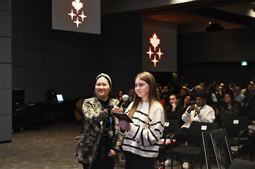 A girl is holding a cue card. A woman holds a microphone for the girl. 
