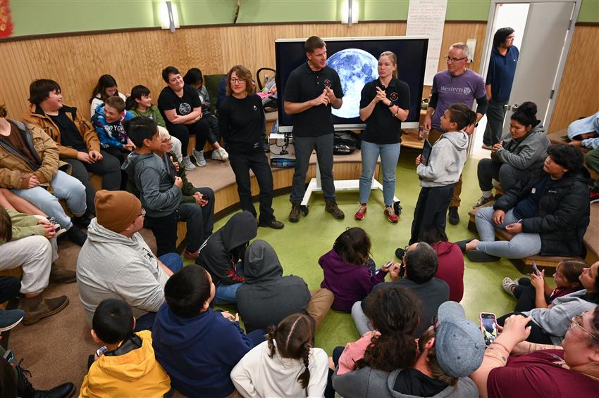 Quatre personnes debout sont entourées d'enfants et d'adultes assis en cercle devant elles.