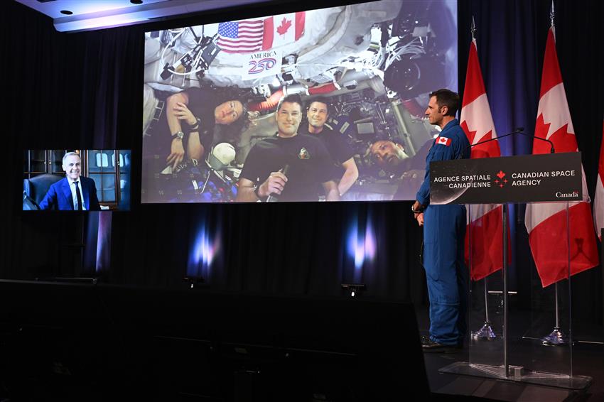 Joshua is standing on stage, looking at the Artemis II crew on a big screen and Mark Carney on a smaller screen.